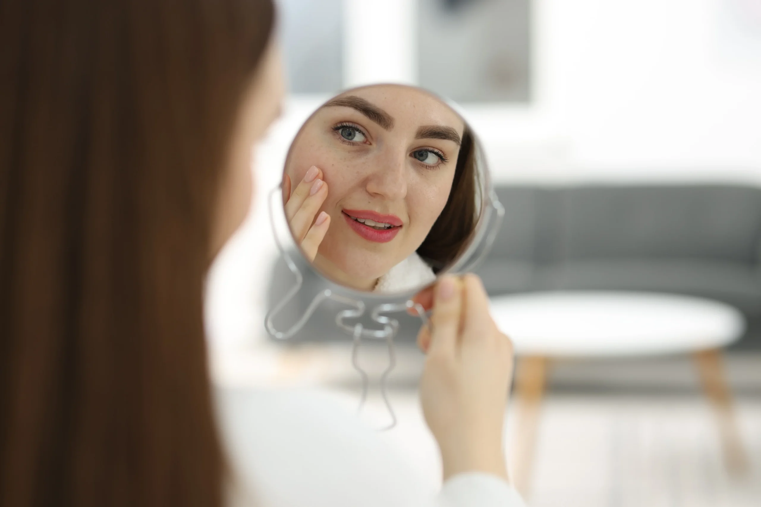 woman examines face with mirror on white - How Can You Clean Clogged Pores From Sunscreen and Sweat?